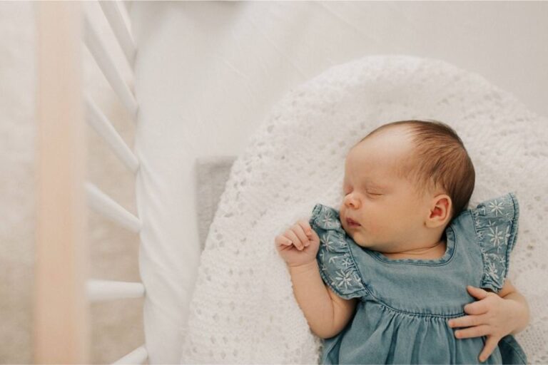 image of newborn baby lying in her white cot wearing a denim blue dress.