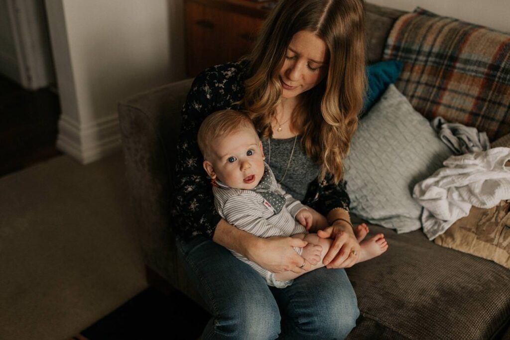 Mother looking down at her baby who's sitting on her lap at home in Plymouth during an intimate motherhood session