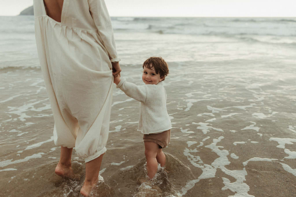 mother holds toddlers hand as they paddle in the sea during lifestyle family photography session