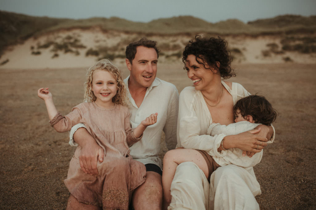 family of four sit on beach laughing with each other whilst toddler nurses from mother