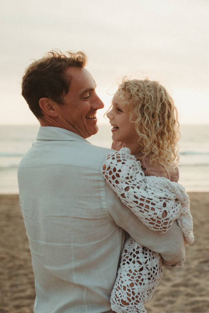 Dad holds daughter snuggled up in blanket both laughing on the beach