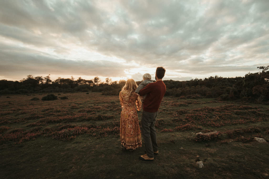 Back of couple and child looking off into moody skies sunset family photographer devon shoot
