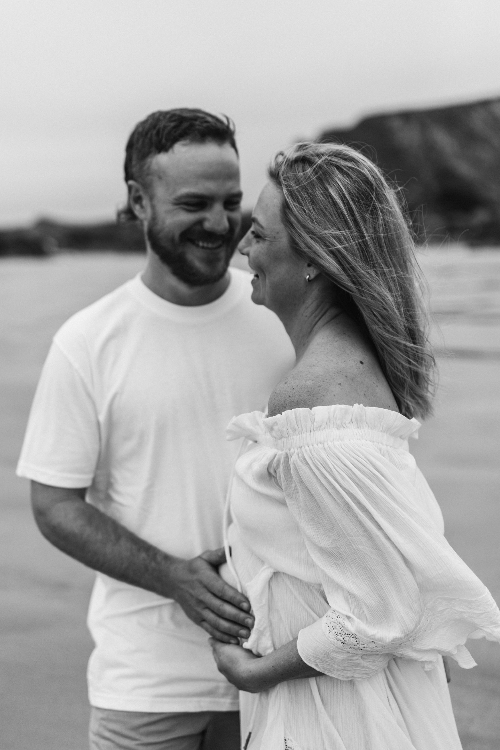 Couple face each other laughing whilst man lays his hands on woman's pregnant stomach maternity photoshoot Cornwall