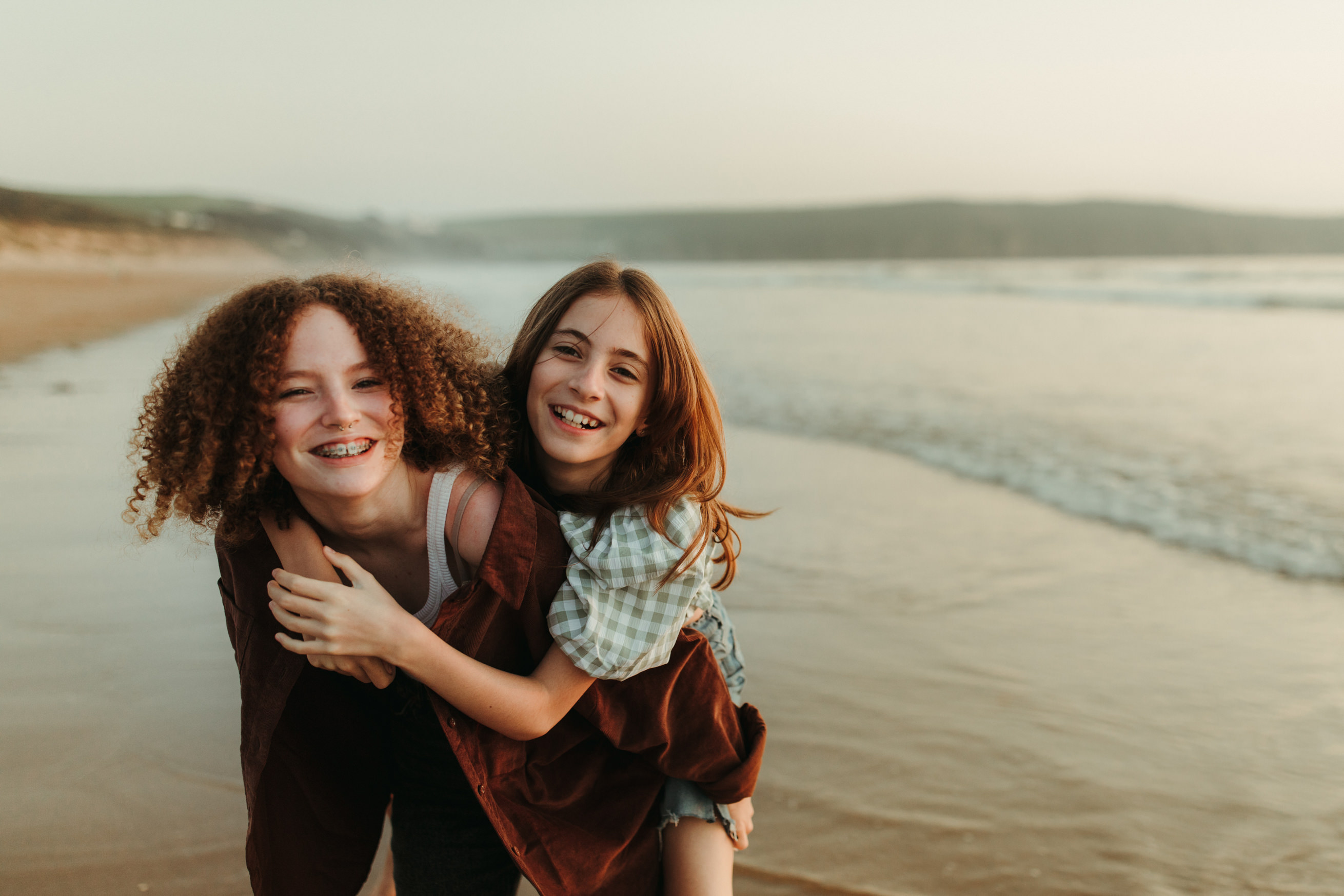 An image of teenage sisters giving piggy-back rides in the sea, during their family photography session with Melanie Grace