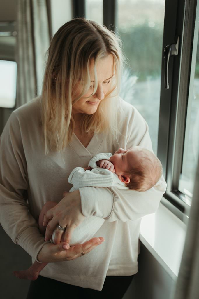 Mum standing next to a window, holding her baby daughter and gazing down at her. Wadebridge Newborn Photographer.