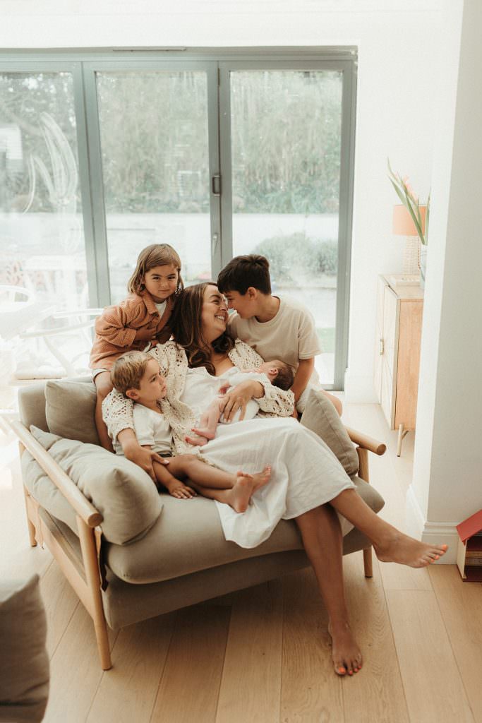 a mother holding her newborn baby wilst being cuddled by her three older boys during a relaxed photography session with Melanie Grace, Cornwall newborn photographer