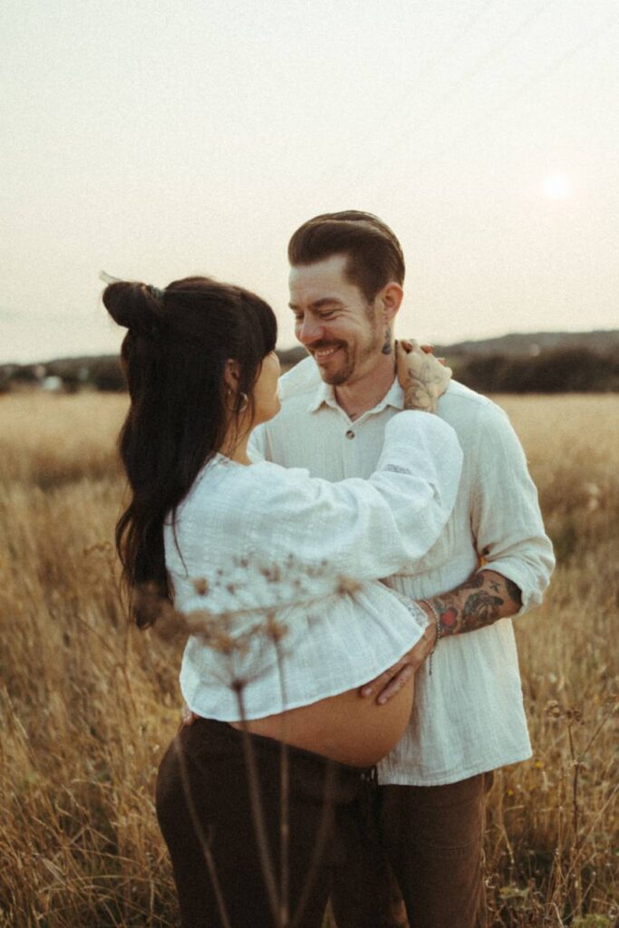 natural maternity portrait of couple standing in long grass holding each other and laughing during golden hour