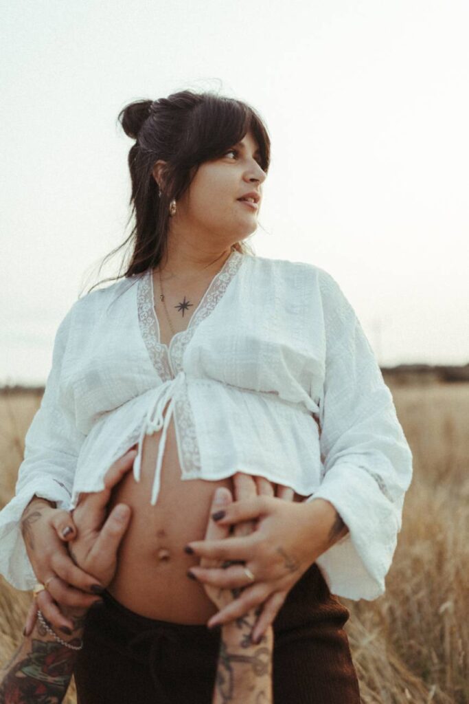 A mother holding her partners hands to her bump and looking into the distance during their relaxed maternity Photography session in Cornwall