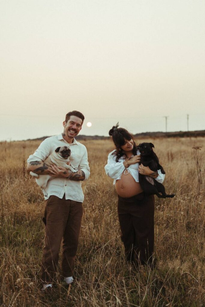 A couple holding pet pug dogs during their sunset maternity photoshoot