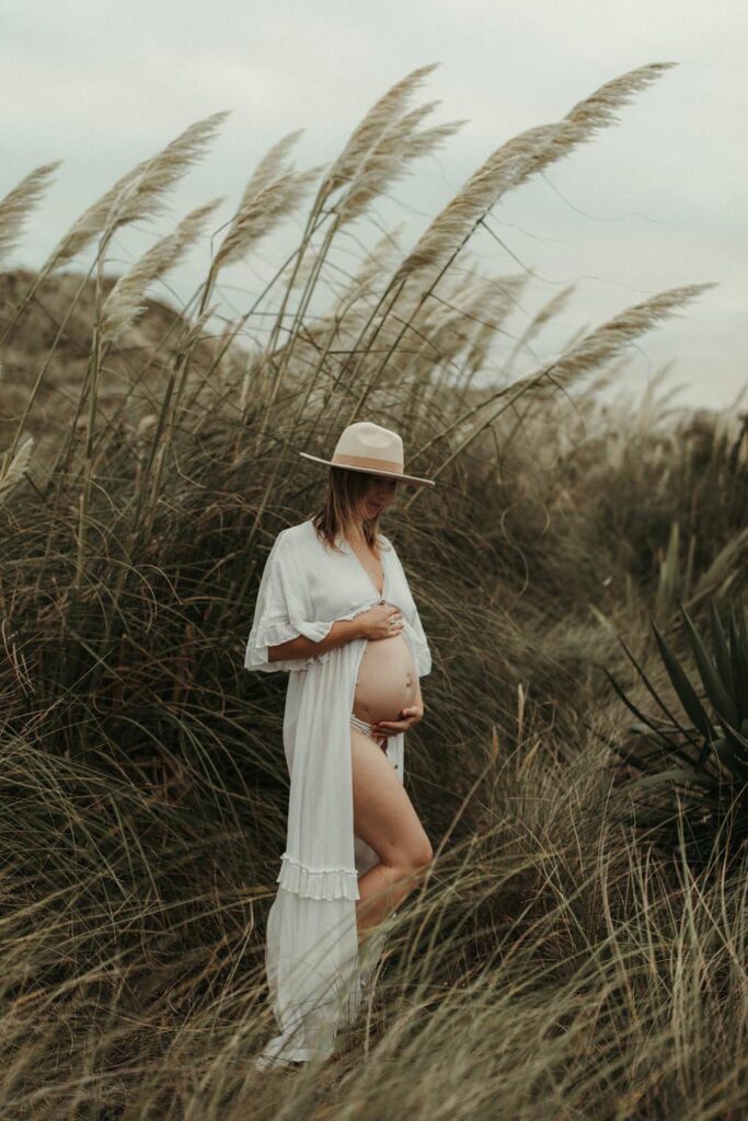 Expectant mother holding her bump surrounded by seagrass and pampas during her maternity photoshoot in Cornwall