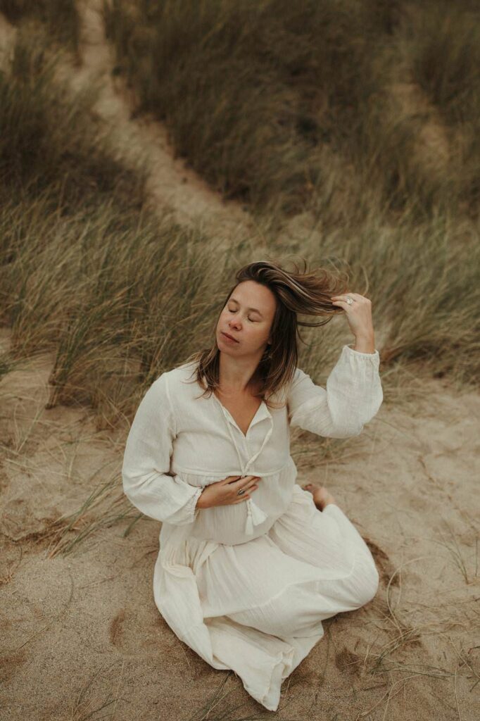 Expectant mother holding her bump and brushing her hair from her face during her maternity photography session in Cornwall