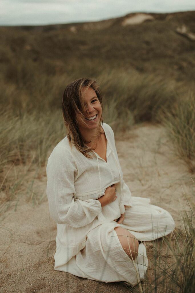 A mother laughing at the camera and holding her baby bump during her maternity photography session in Cornwall