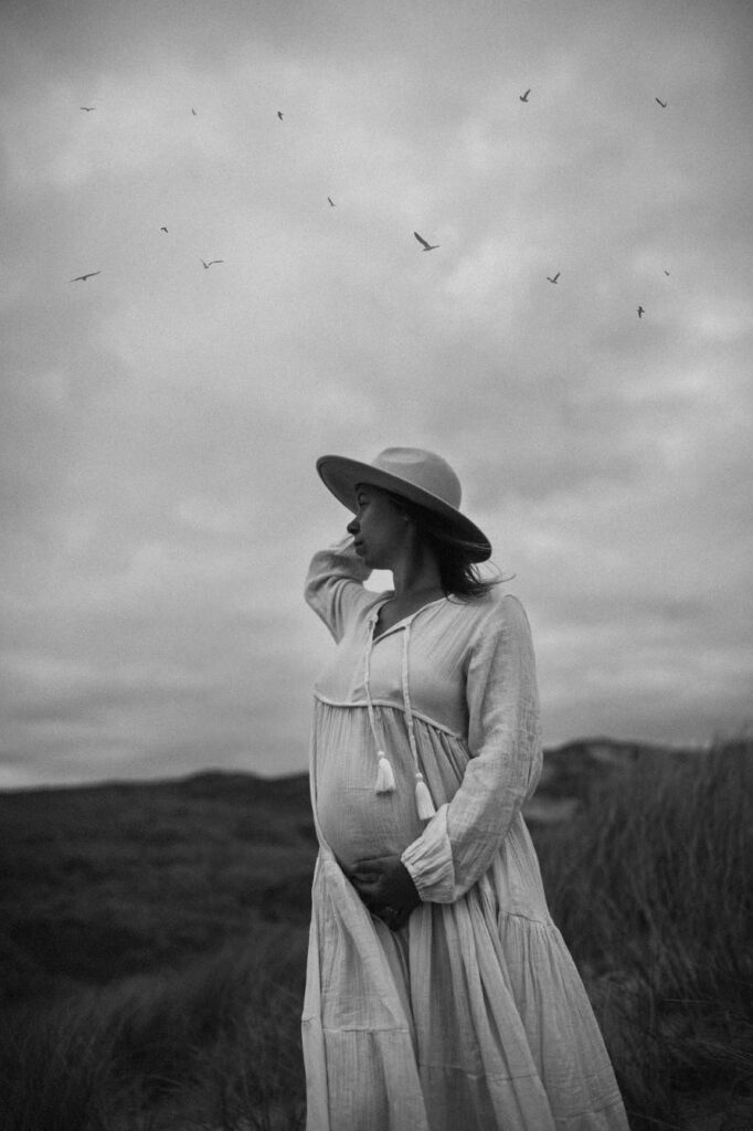 Pregnant woman standing on the beach with flowing dress and birds flying in the sky above her