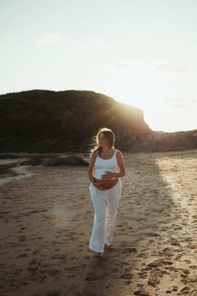 Expectant mother holding her bump by the sea in soft golden light at Constantine Bay in Cornwall
