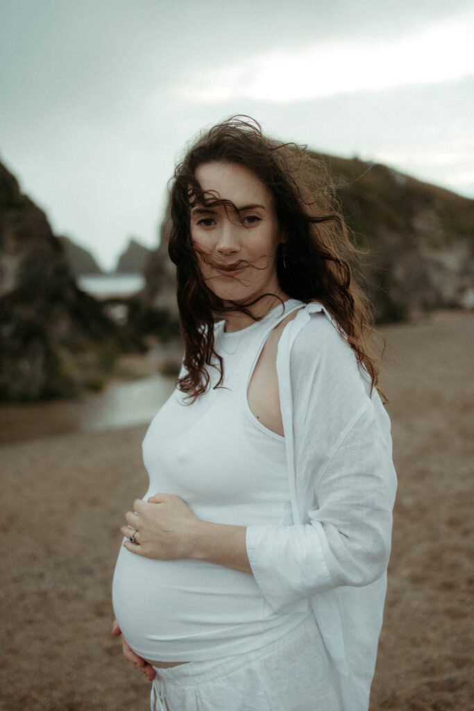 Expectant mother holding her bump while her hair gently blows across her face at Holywell Bay in Cornwall